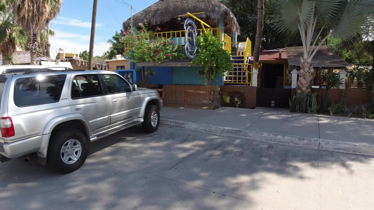 At 0:00 in Los Cabos, Mexico, a silver SUV fills the left foreground beside a thatched-roof building with yellow railings, palm trees, blue sky, and no visible people.