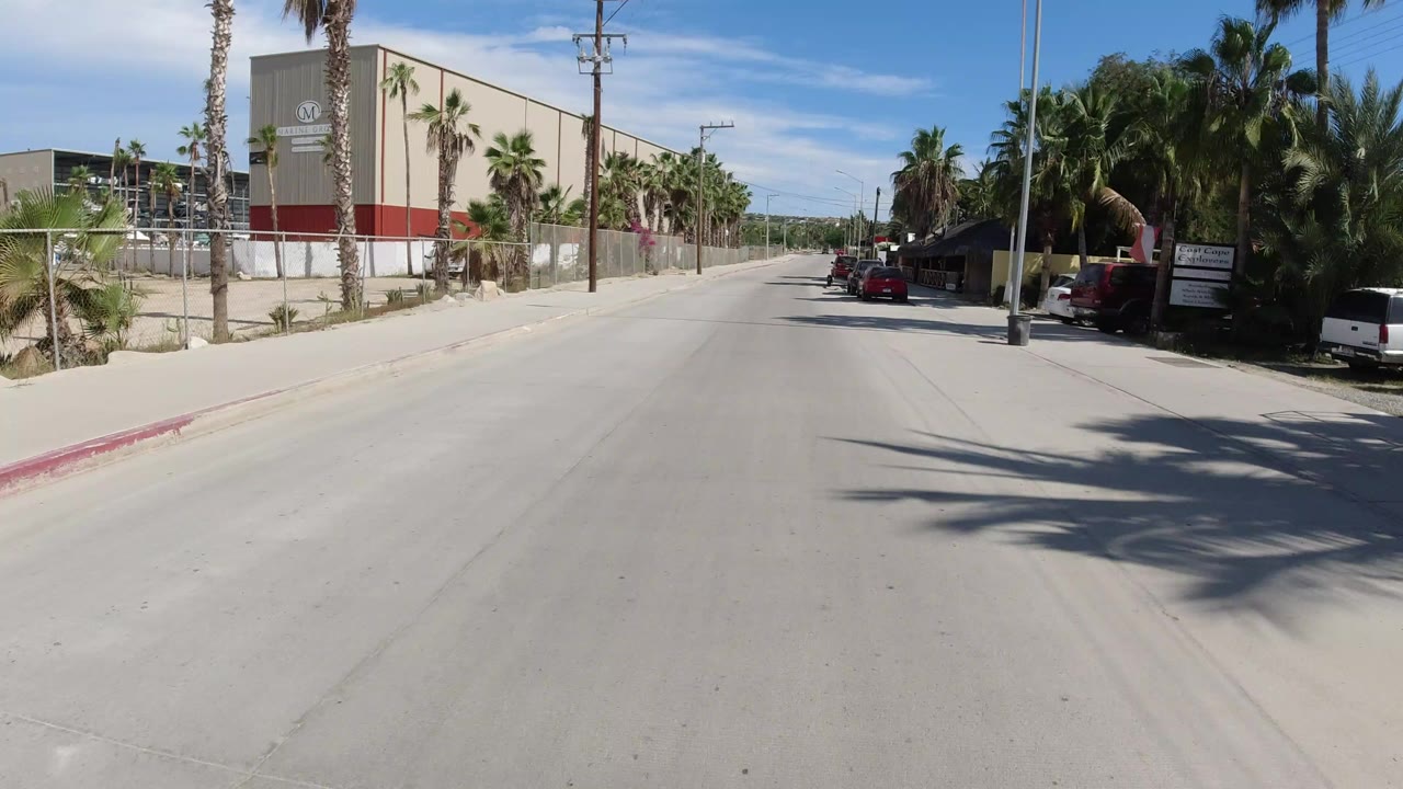 Road-level view of a Los Cabos street with beige buildings behind chain-link fencing on the left, parked cars and palms on the right, blue sky overhead, and no visible people.