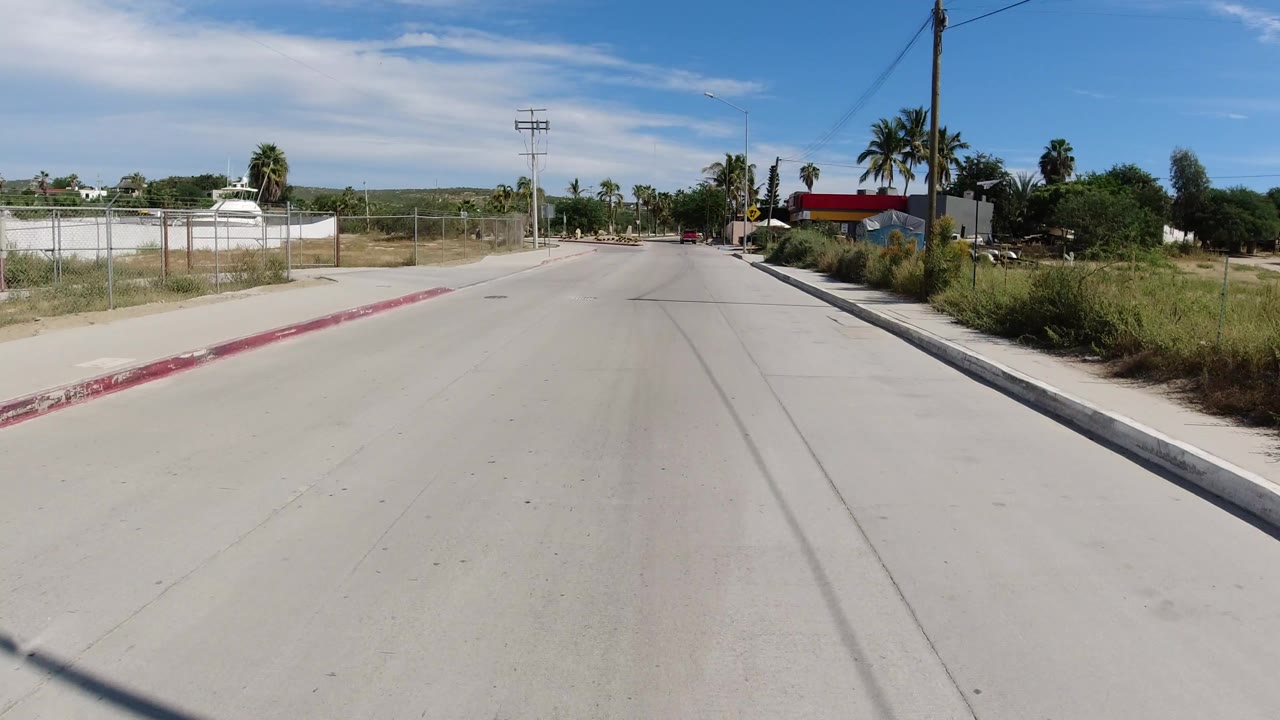 A forward-facing Los Cabos road frame shows fencing and dry roadside lots on the left, palms and low buildings ahead, and no visible people.