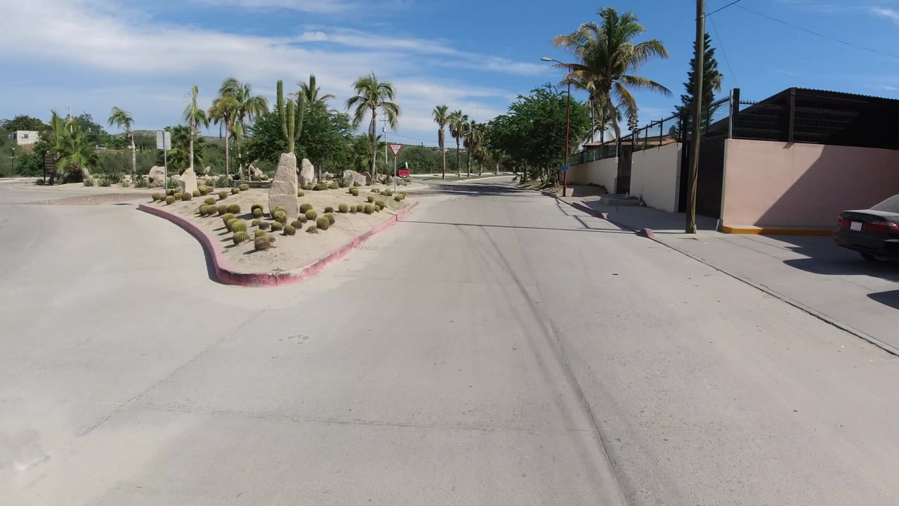 A wide road-level view in Los Cabos, Mexico shows a paved intersection, palm trees, desert landscaping, a walled property, and a dark car at the right edge, with no visible people.