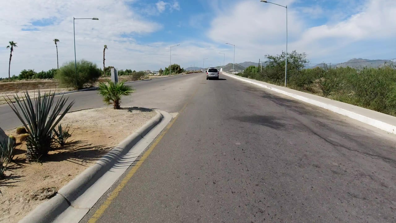 A light-colored Wrangler drives away on a smooth road in Los Cabos, Mexico, with agave-like plants at the curb, streetlights, distant hills, bright clouds, and no visible people.