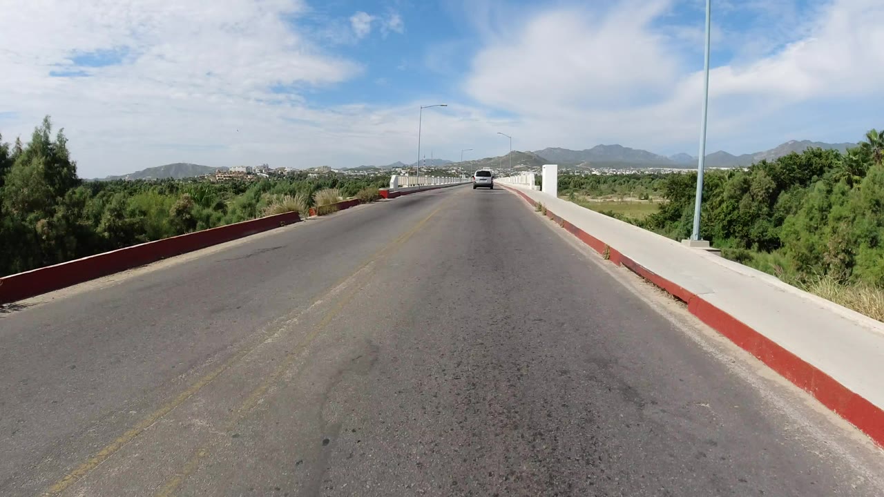 Forward view along a bridge road in Los Cabos, Mexico, with red curb edges, green roadside vegetation, distant hills, a small vehicle ahead, and no visible people.