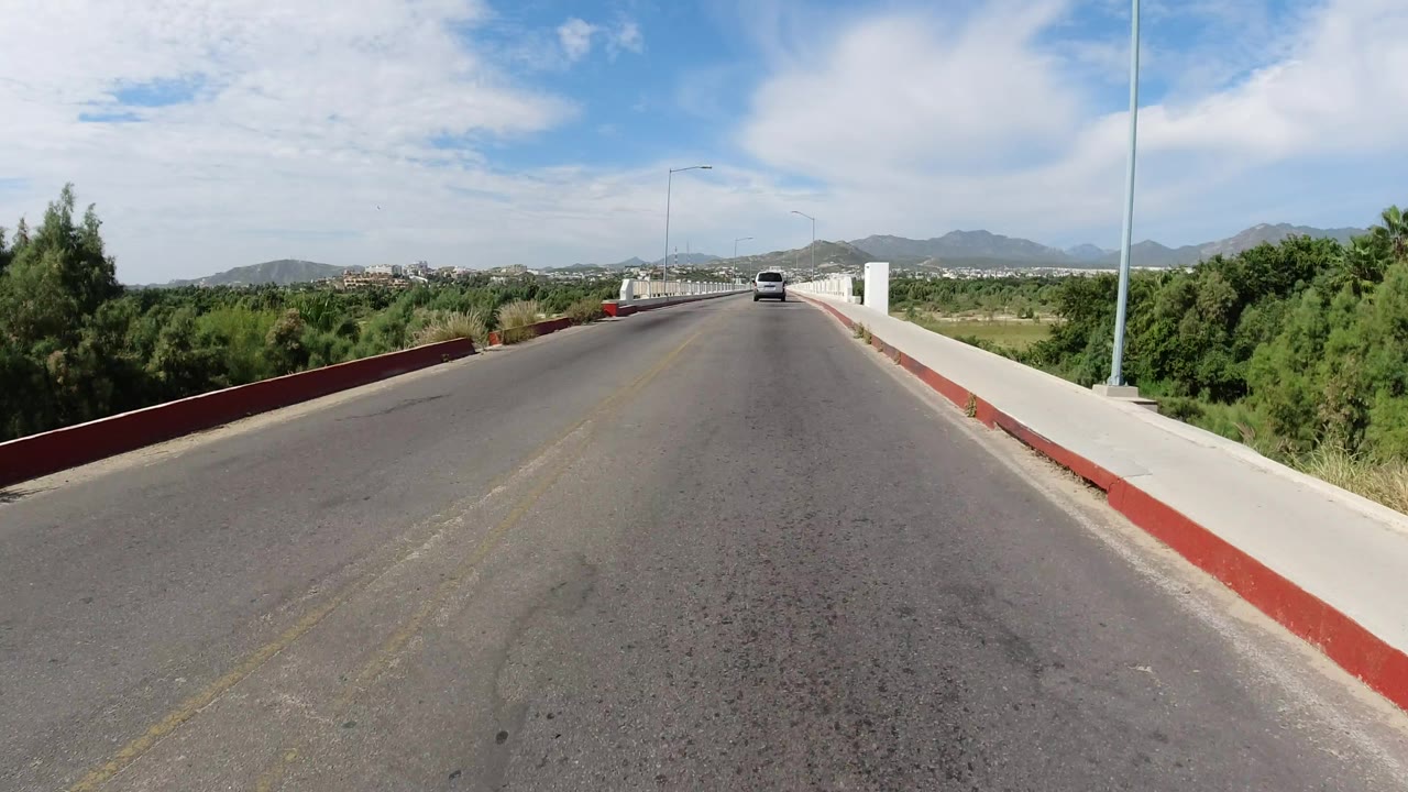Centered Los Cabos bridge roadway view with white sidewalls, a streetlight pole, low green vegetation on both sides, mountains on the horizon, and no visible people.