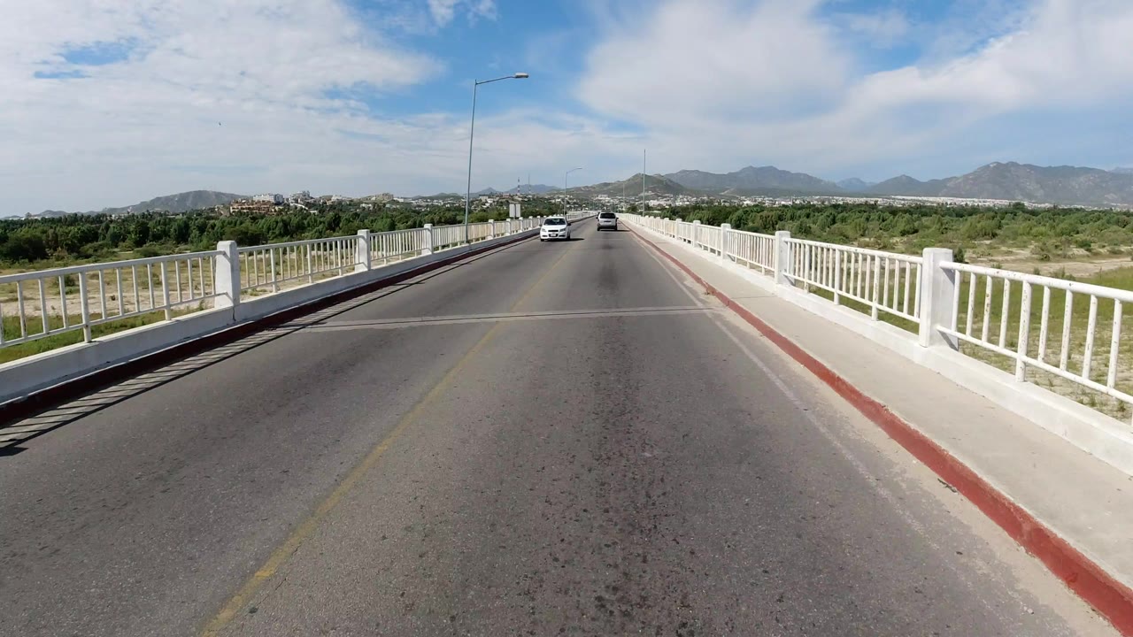 A sunlit road bridge in Los Cabos, Mexico stretches ahead between white railings, with distant cars, low hills, blue sky, and no visible people.
