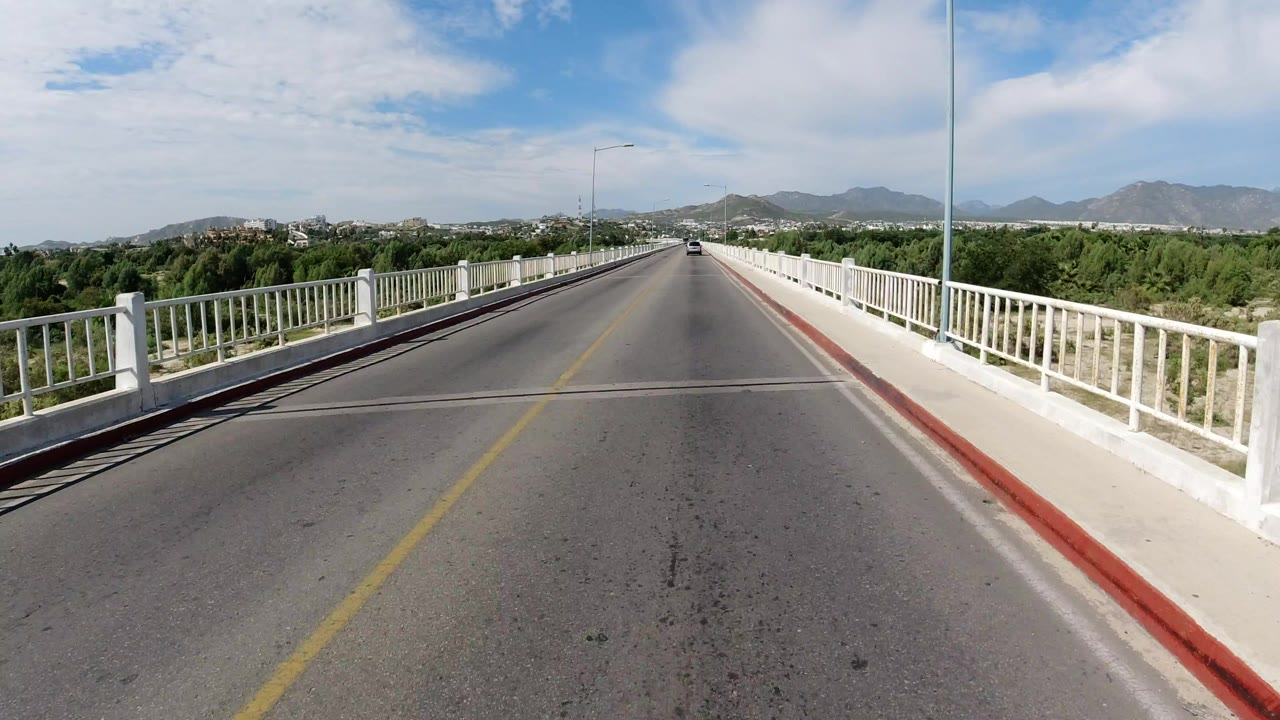 A forward view down a sunny bridge road in Los Cabos, Mexico, with white railings, green trees, distant buildings, low mountains, and no visible people.