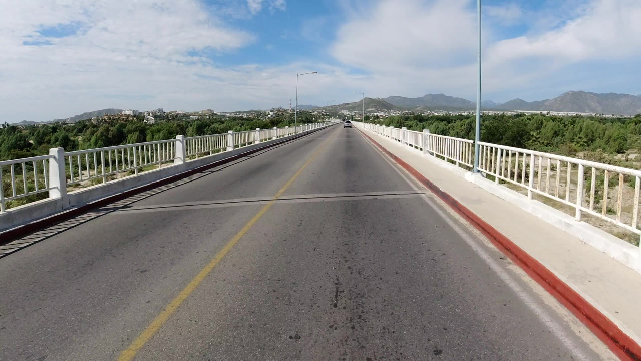 The Los Cabos bridge road remains centered, with the right railing close to camera, trees below the bridge, hills ahead, and no visible people.