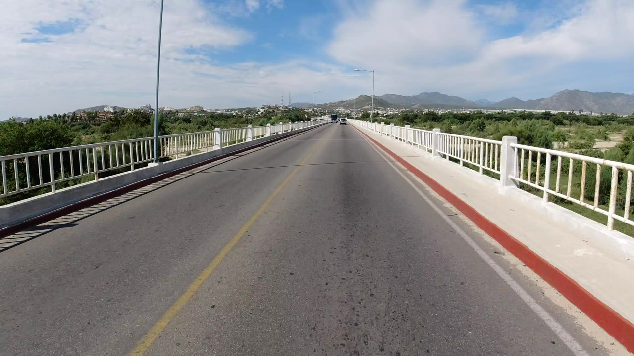 Start of a Los Cabos, Mexico bridge crossing with white railings on both sides, a red curb edge, green vegetation, distant hills, bright clouds, and no visible people.