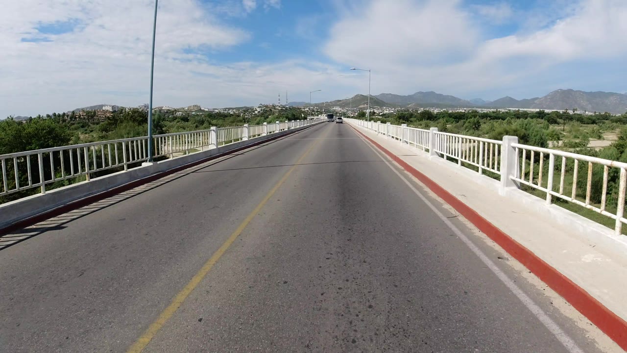 Early frame of the Los Cabos bridge approach with the road centered, white railings tapering toward the horizon, roadside greenery, distant hills, and no visible people.