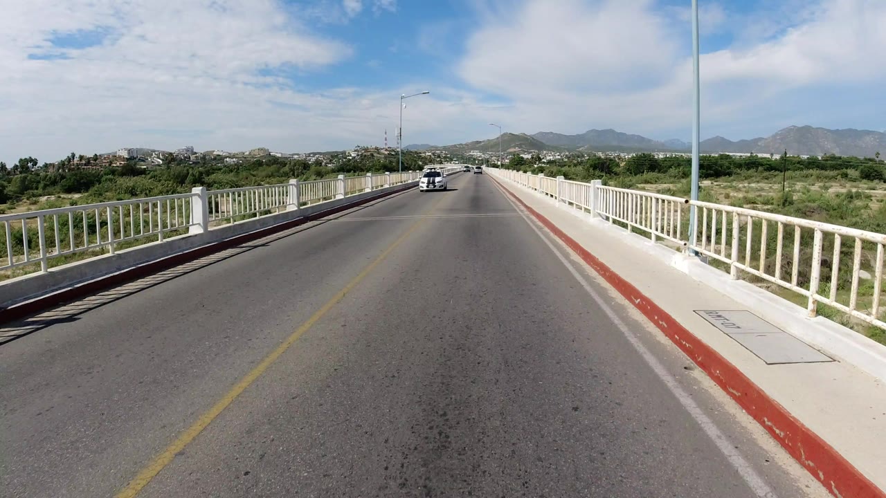 Los Cabos roadway continues across the bridge, with the white car farther along the lane, white guardrails, low green vegetation, mountains on the horizon, and no visible people.