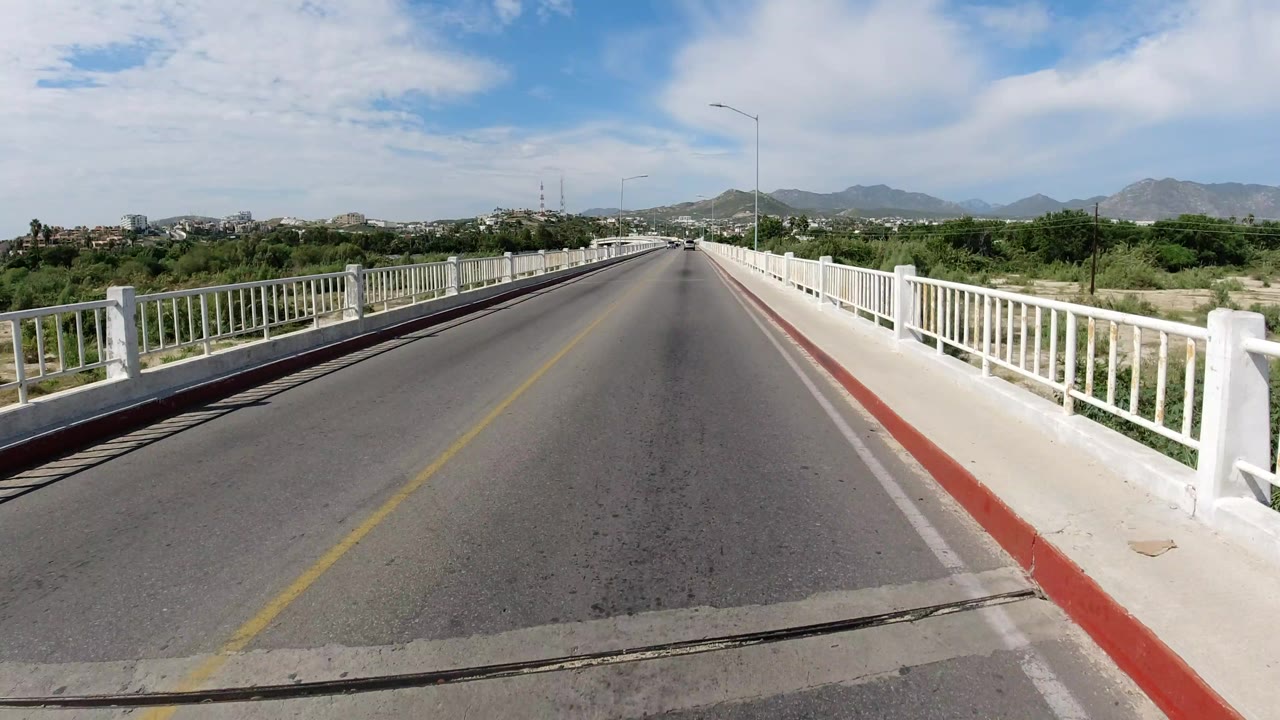 The second frame keeps the Los Cabos bridge centered, showing white railings, a red curb edge, open asphalt, roadside greenery, distant hills, and no visible people.