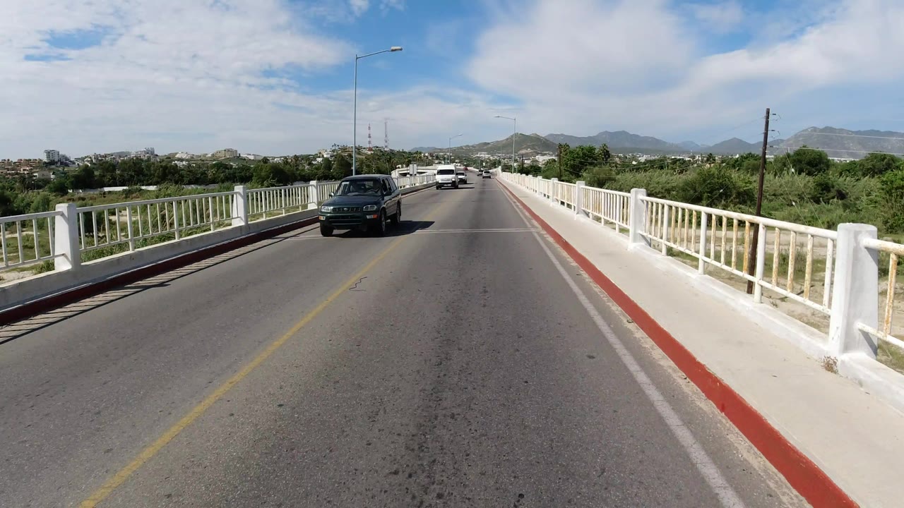A Los Cabos bridge roadway seen from the lane center, with a dark car ahead, white railings on both sides, green roadside growth, distant hills, and no visible people.