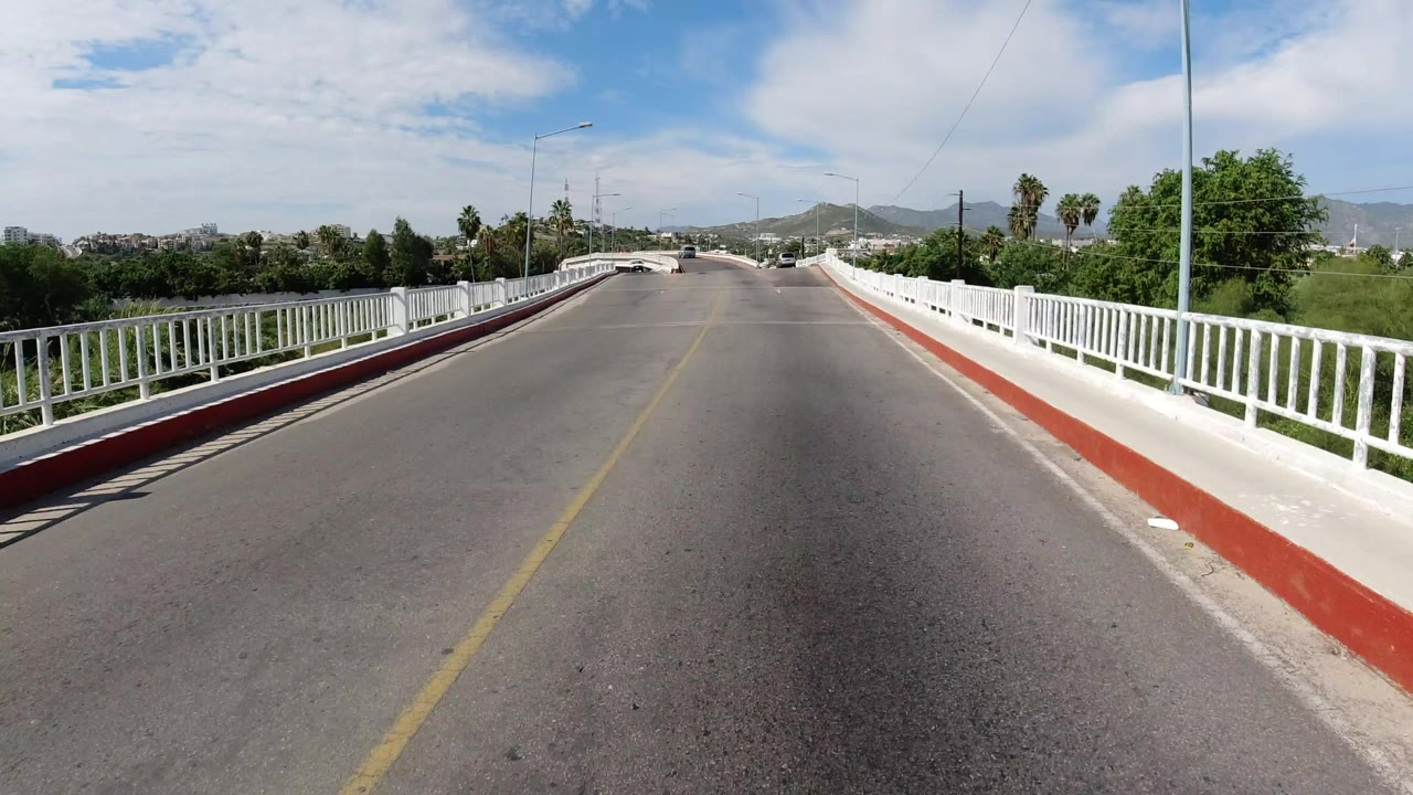 The Los Cabos bridge road continues under bright daylight, with the right railing prominent, palms beyond the rail, hills in the distance, and no visible people.