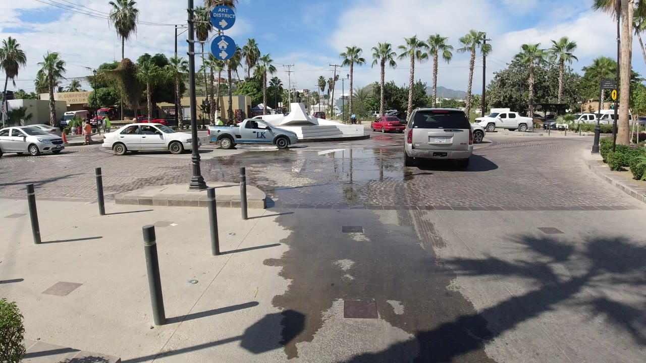 Los Cabos roundabout seen from a sidewalk with black bollards, wet cobblestones, palm trees, cars, street signs, and small pedestrians near the left background.