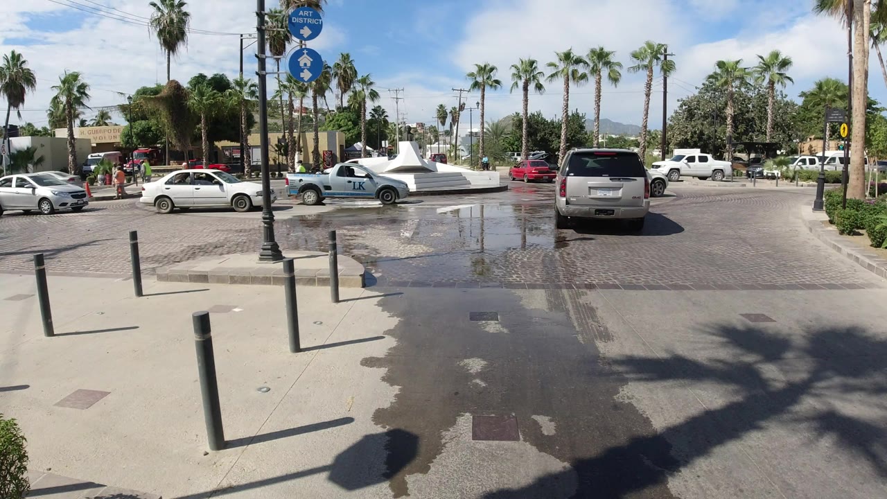 Los Cabos street scene with a white car and light blue pickup near the roundabout, wet road reflections, palms, signs, and distant pedestrians.