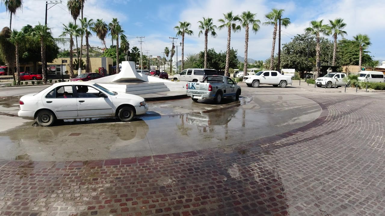 A white sedan sits left of frame while a light blue pickup marked on the tailgate passes through the wet Los Cabos roundabout, with palm trees and no visible people.