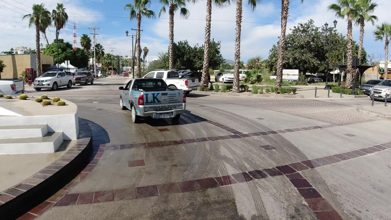 Los Cabos street view one second into the clip, with the camera following a white pickup around curved pavement, palms, a low wall, and no clearly visible people.
