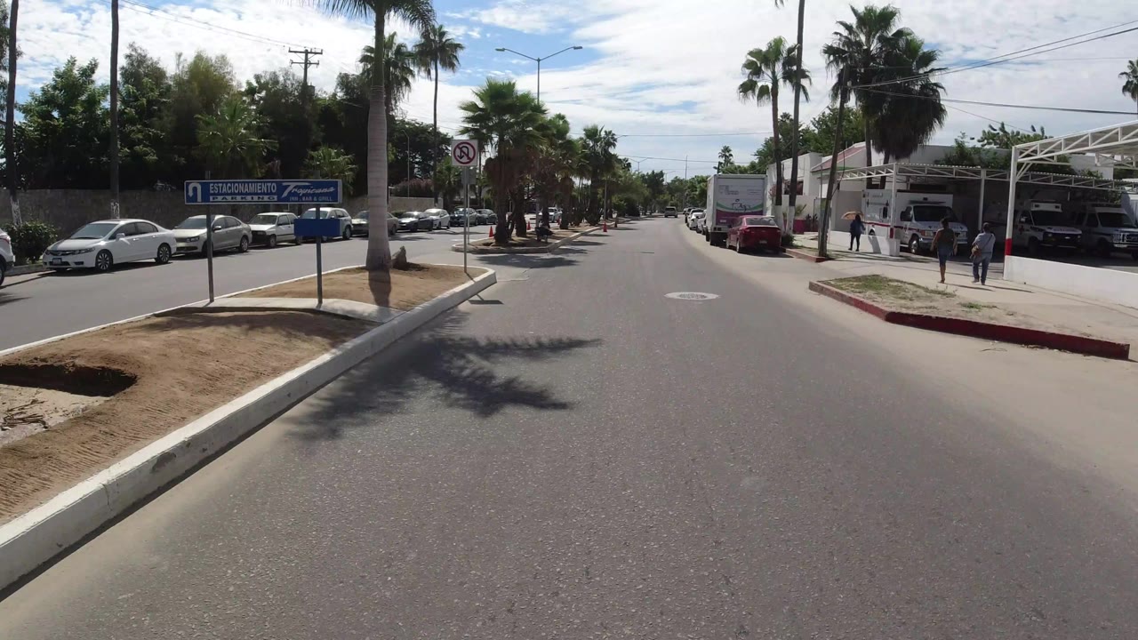 Los Cabos street view one second in, showing the median tip, a no U-turn sign, palm trees, parked cars, and small distant pedestrians near the right-side buildings.