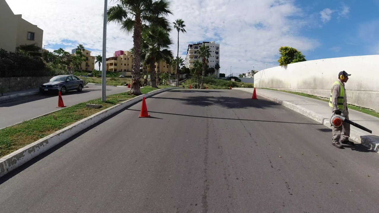 Los Cabos road scene with a worker at the right curb, orange cones marking the street, palm trees in the median area, and apartment buildings beyond the landscaped slope.