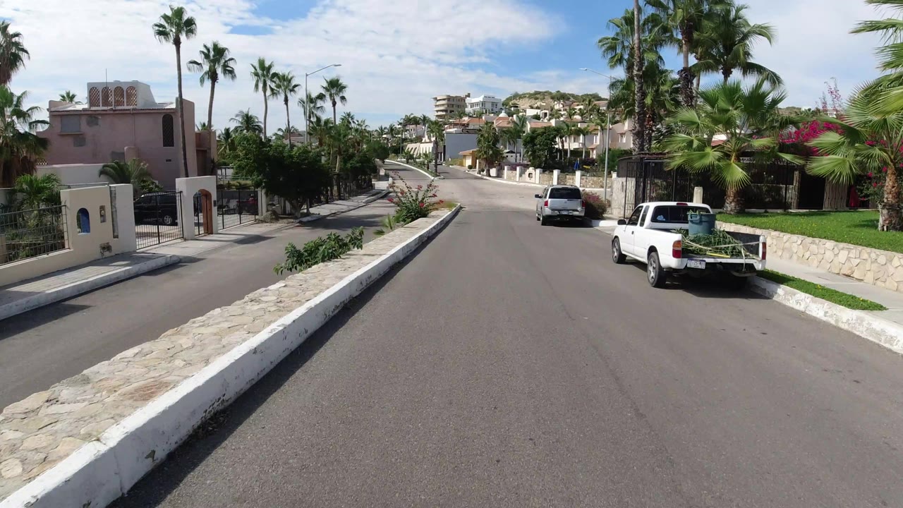 Los Cabos street view slightly farther along the road, with a stone curb median, palm landscaping, a white pickup at right, hillside homes ahead, and no visible people.