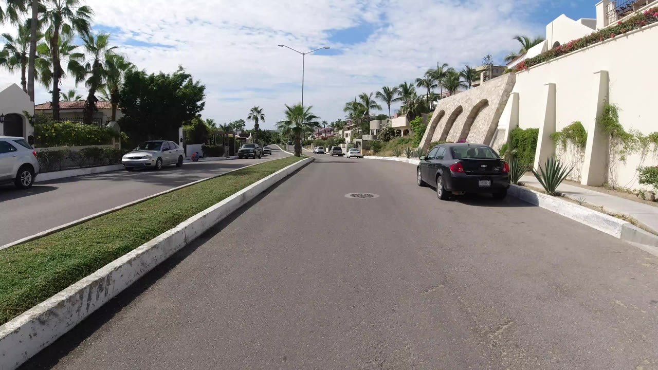 Opening view of a Los Cabos residential street with a grass median, palm trees, cream walls, parked cars, and no visible people.