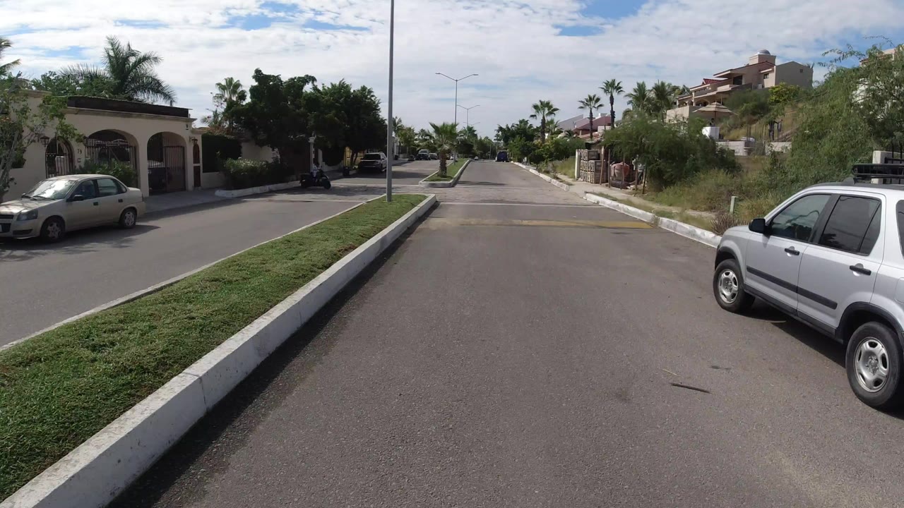 Opening view of a Los Cabos residential road with a raised grass median, parked cars, palms, walled homes, and no visible people.
