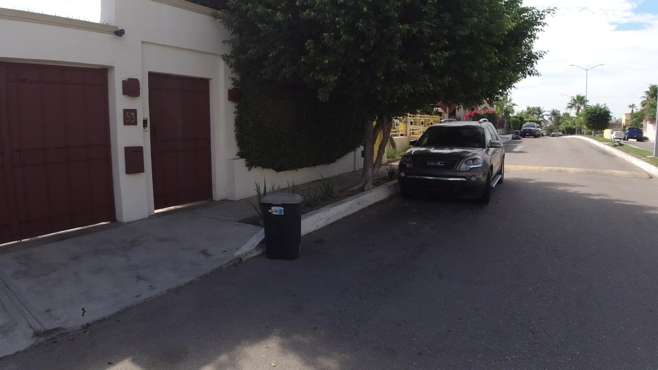 Los Cabos curbside frame looking past red garage doors and a white wall toward a dark parked SUV, trees, distant cars, and no visible people.