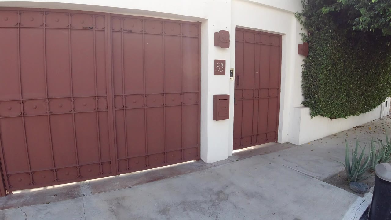 Closed red metal gate in a white wall beside a hedge and sidewalk.