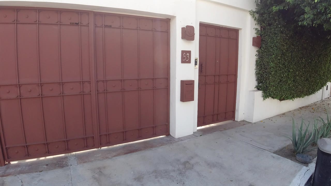 Red gate remains closed in the Los Cabos residential wall, with the side door, address plaque, hedge, agave plants, and empty concrete pavement visible.