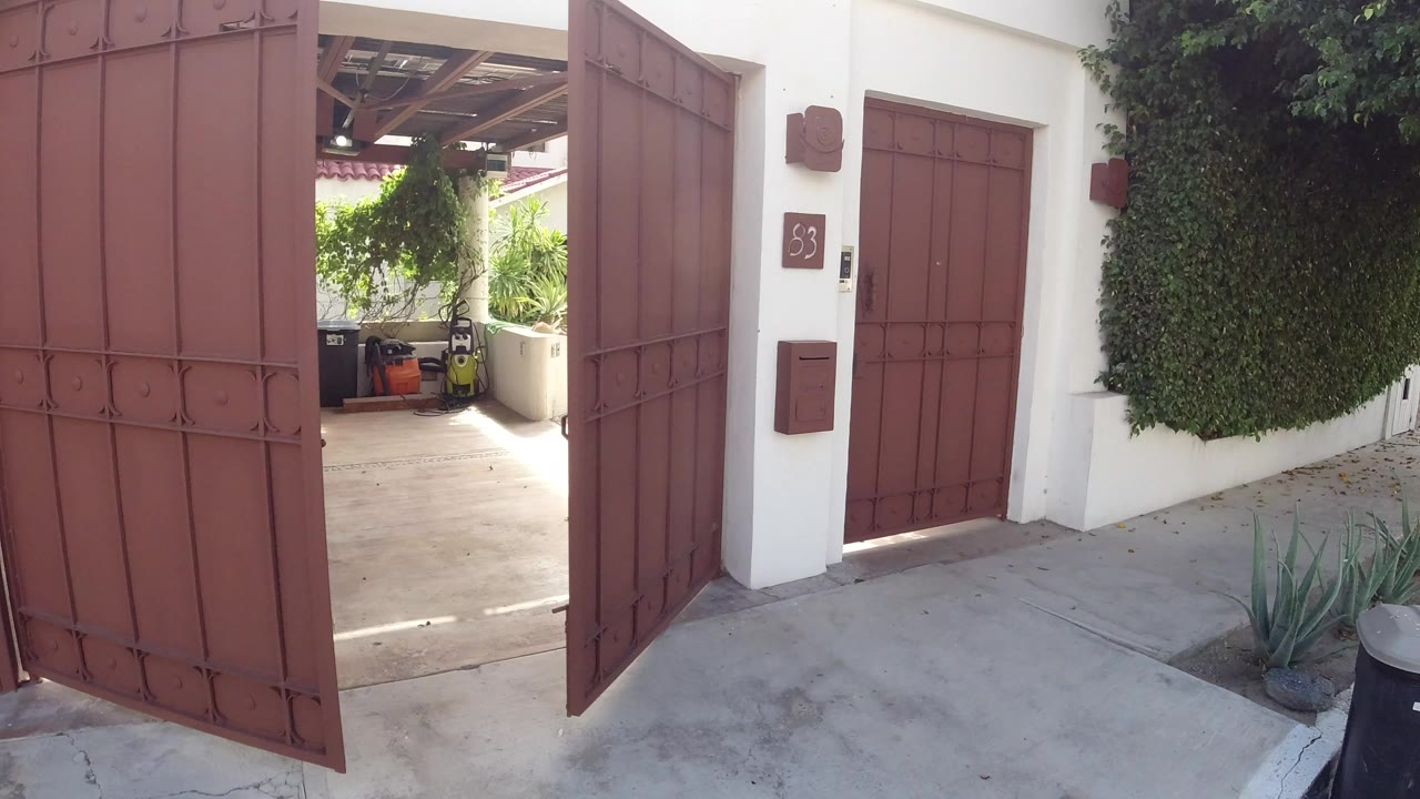 Opening frame in Los Cabos, Mexico, showing a red metal gate partly open beside a white stucco wall, a red door, greenery, and no visible people.
