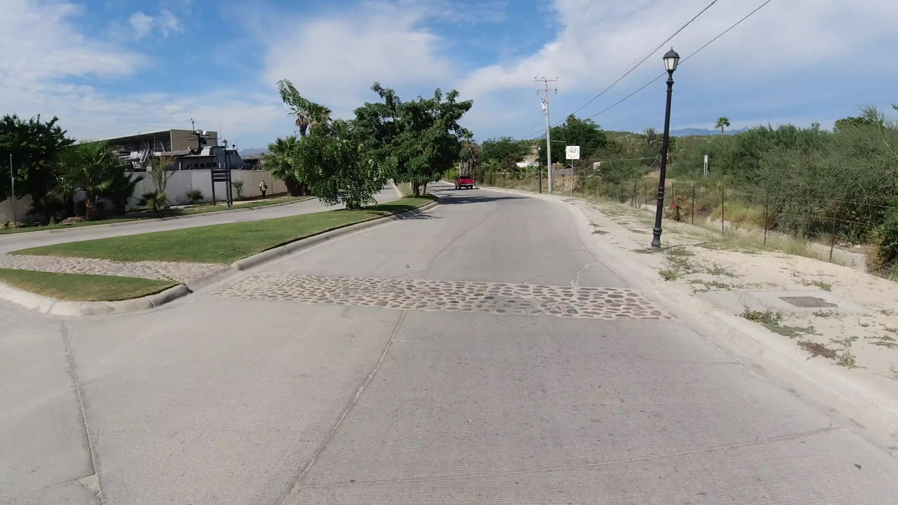 Los Cabos street view with a red vehicle in the distance, a speed sign on the right, dense trees on the left, and no visible people.