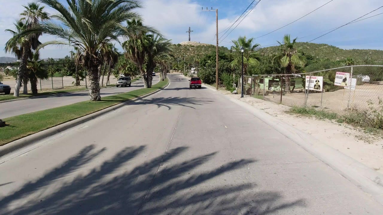 Start of a Los Cabos road view with palm trees on both sides, a red vehicle ahead, fenced roadside lots, distant hills, bright sun, and no visible people.