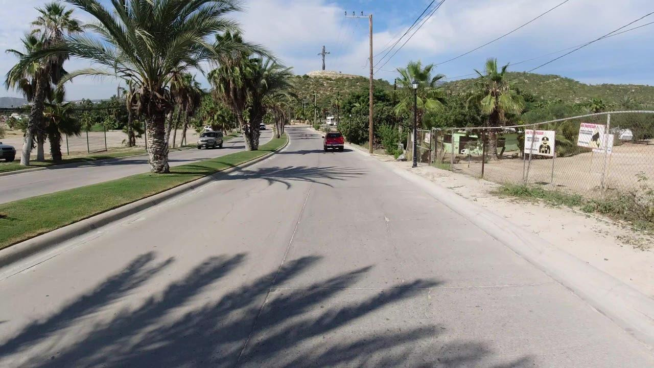 Los Cabos road scene one second in, with a red vehicle farther ahead, palms casting shadows across the lane, a grassy divider, roadside fencing, and no visible people.