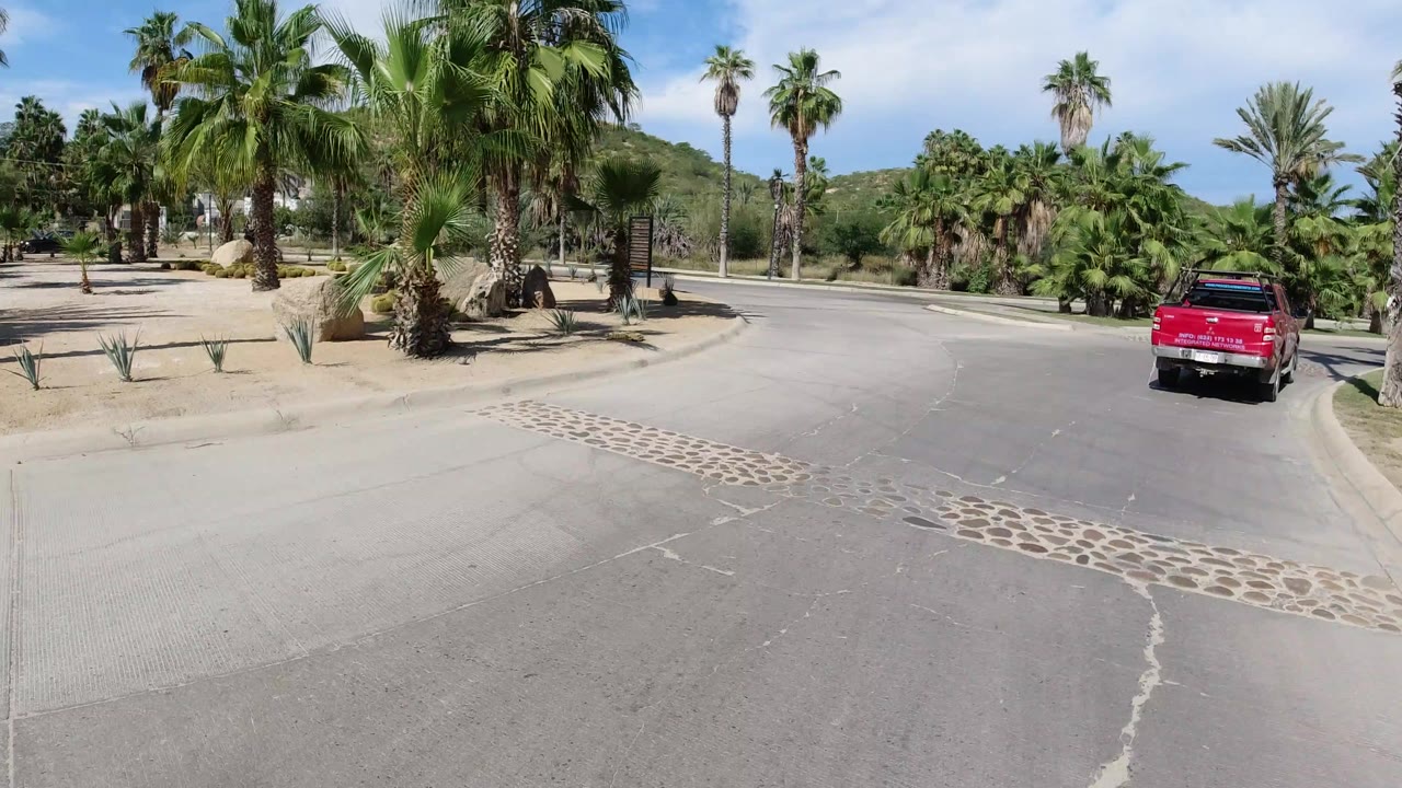 A red pickup drives along a broad paved road in Los Cabos, Mexico, beside curbs, palms, desert plants, and low hills, with no visible people.