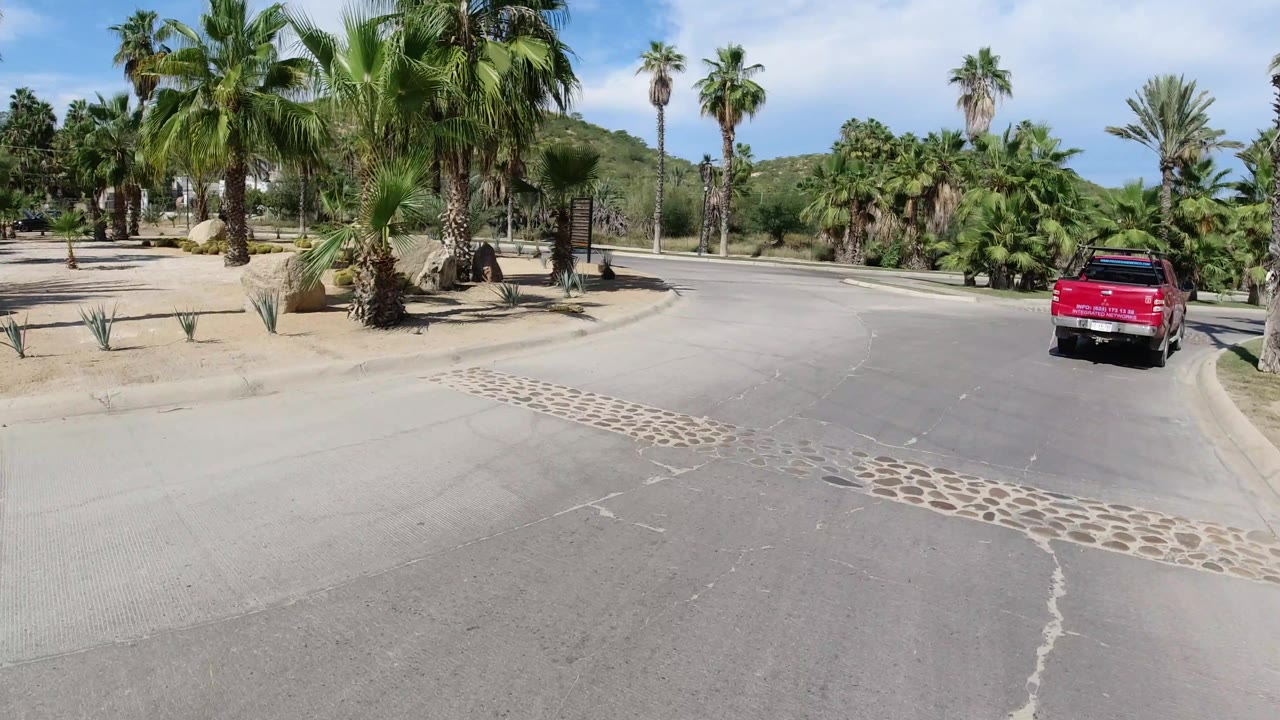 The red pickup continues along the curved Los Cabos road with palms, landscaped islands, pale pavement, and low green hills in the background, with no visible people.