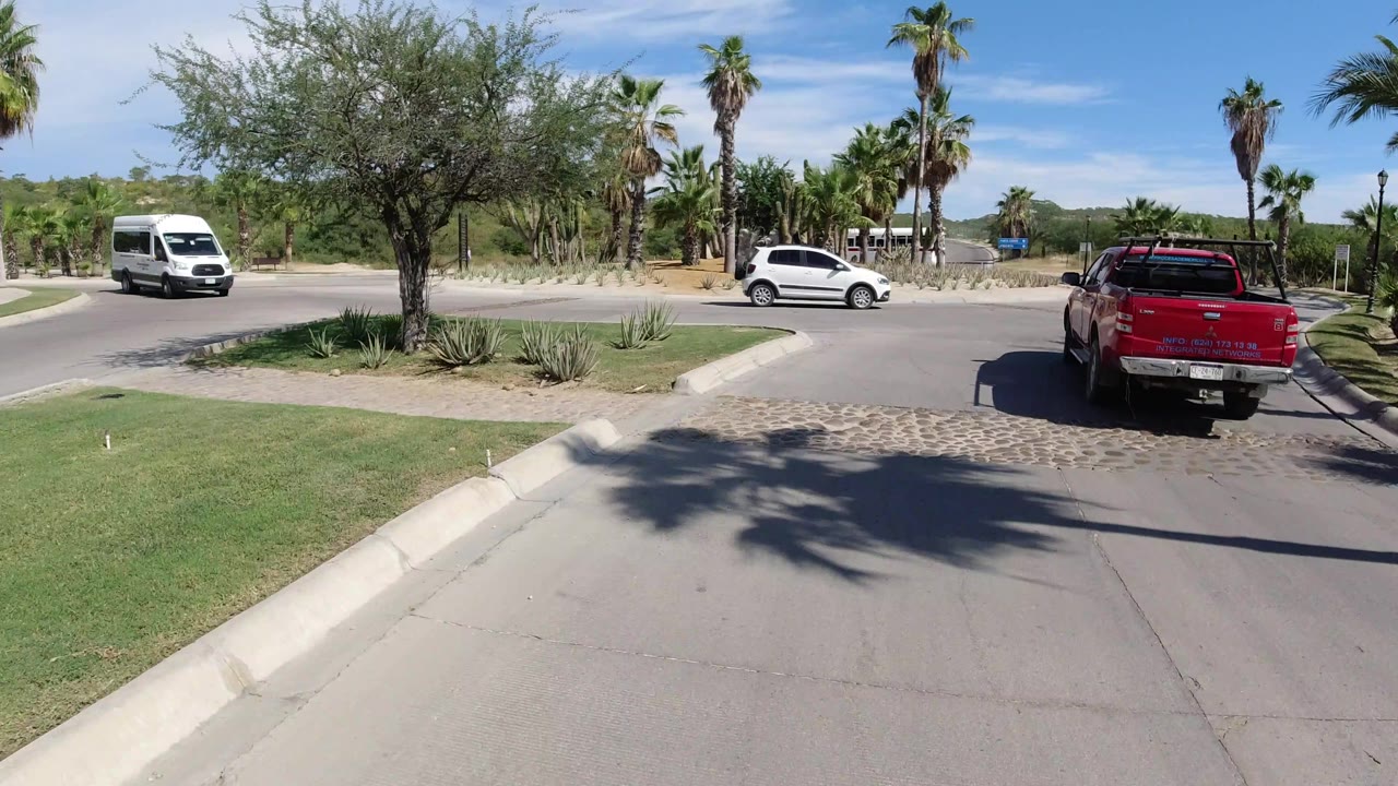 Los Cabos roundabout view with the red pickup farther ahead on the right, a white van entering from the left, a silver car crossing midframe, palm trees, agave beds, and no visible people.