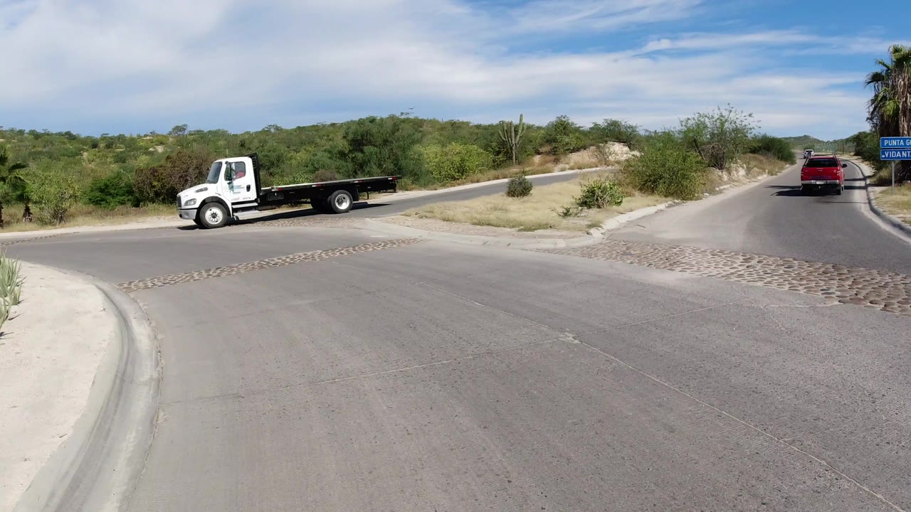 Wide Los Cabos road view with a white flatbed truck at left, a red SUV farther ahead, dry shrubs, palms, low hills, and no visible people.
