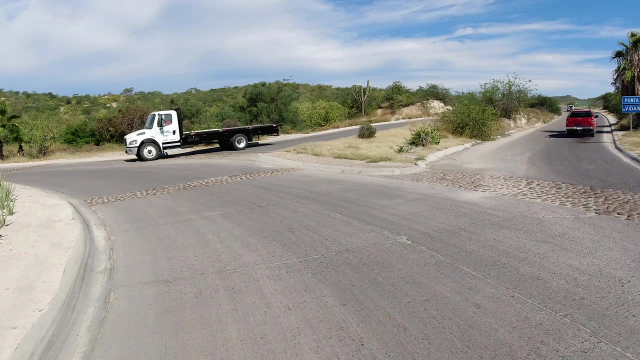 Los Cabos street scene with the white flatbed truck moving through the left side of the frame, a red SUV up the road, dry vegetation, palms, and no visible people.