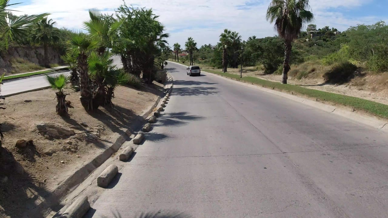 The Wrangler continues along the Los Cabos roadway with palms on both sides, a dry slope to the left, open greenery to the right, and no visible people.
