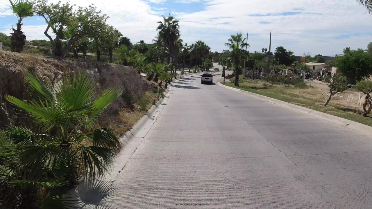 The Los Cabos road continues past palms and a rocky dirt embankment, with the van centered farther down the lane and no clearly visible people.