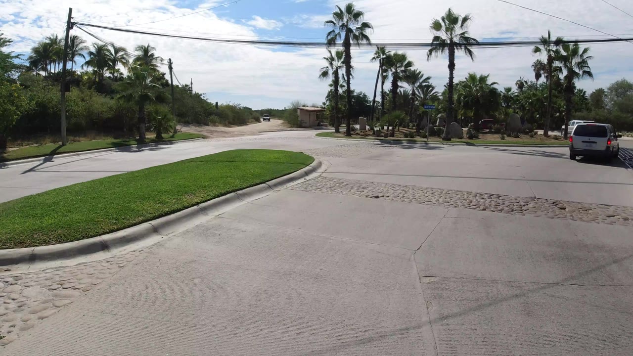 Los Cabos roadway curves past a grassy island, palms, desert shrubs, and roadside landscaping under bright sun, with a white van farther right and no visible people.