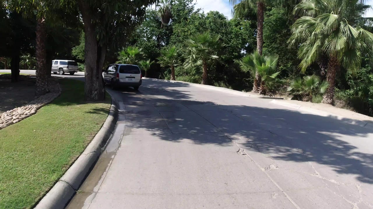 One second into the Los Cabos road clip, the Jeep remains ahead near the left side of the lane as palms, lawn, and dark tree shade frame the route, with no visible people.