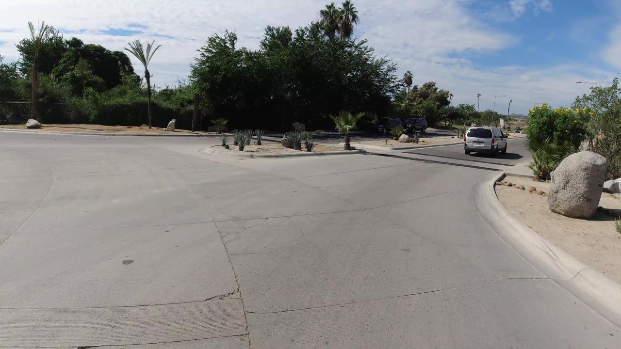 Los Cabos vehicle viewpoint beside a curved curb, with roadside rocks, desert plants, parked cars, a white car ahead, bright sun, and no visible people.