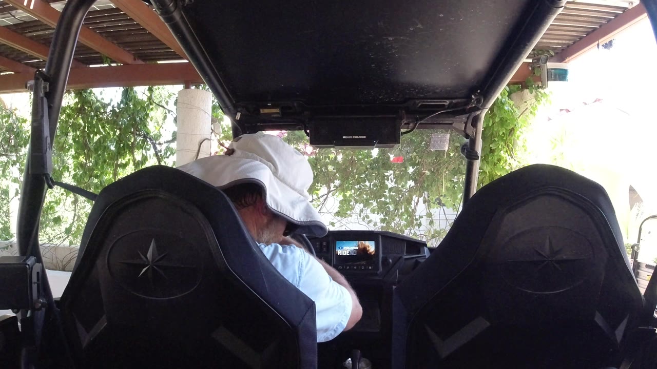 The man remains leaned forward inside the Los Cabos vehicle, with the dashboard screen visible ahead and bright foliage outside the open side.