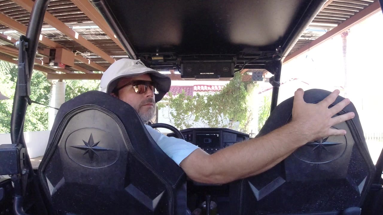Adult man in a white bucket hat and sunglasses sits inside a black golf cart in Los Cabos, Mexico, with his hand resting on the seat back and bright exterior light beyond the vehicle.