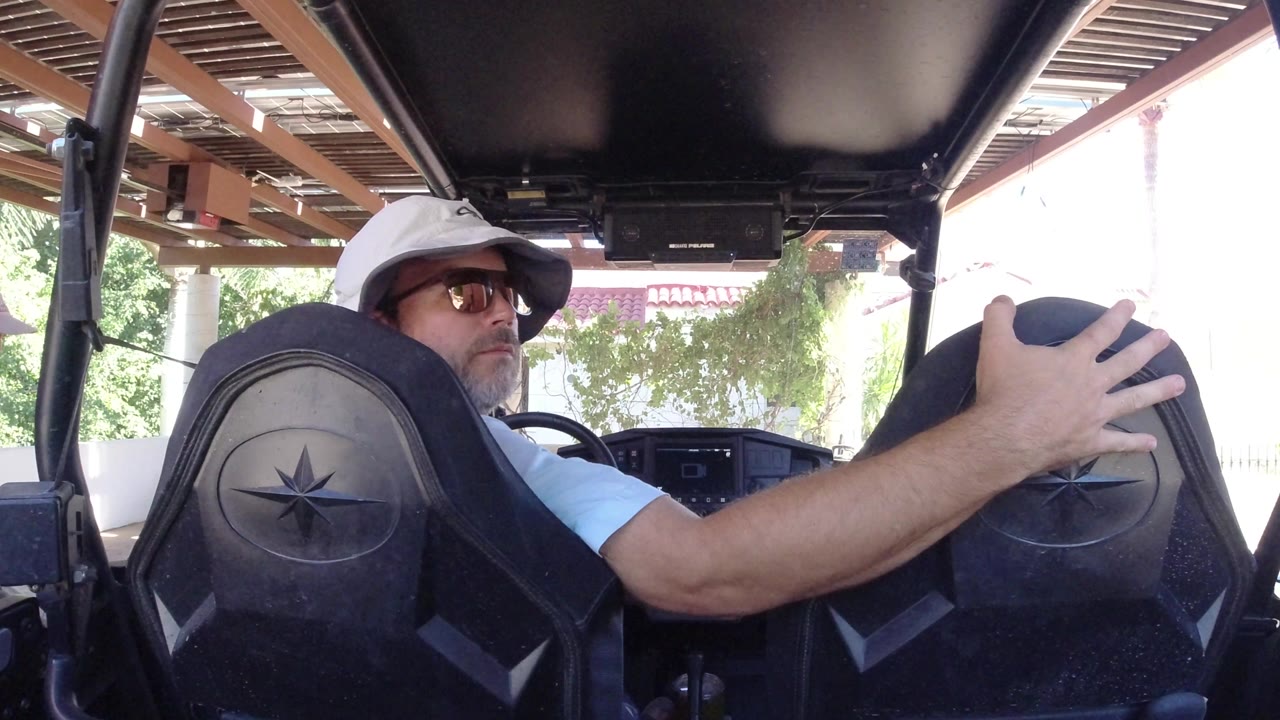 The driver looks back over his shoulder from inside the golf cart, with the cart roof overhead, black seats in the foreground, and sunlit greenery outside in Los Cabos, Mexico.