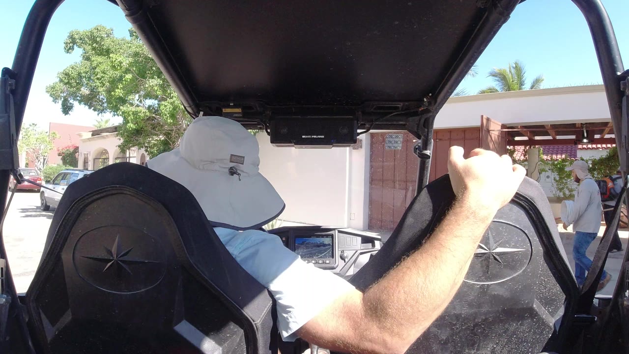 The driver remains framed between black buggy seats, with his right arm raised and pedestrians visible near the building entrance in Los Cabos, Mexico.