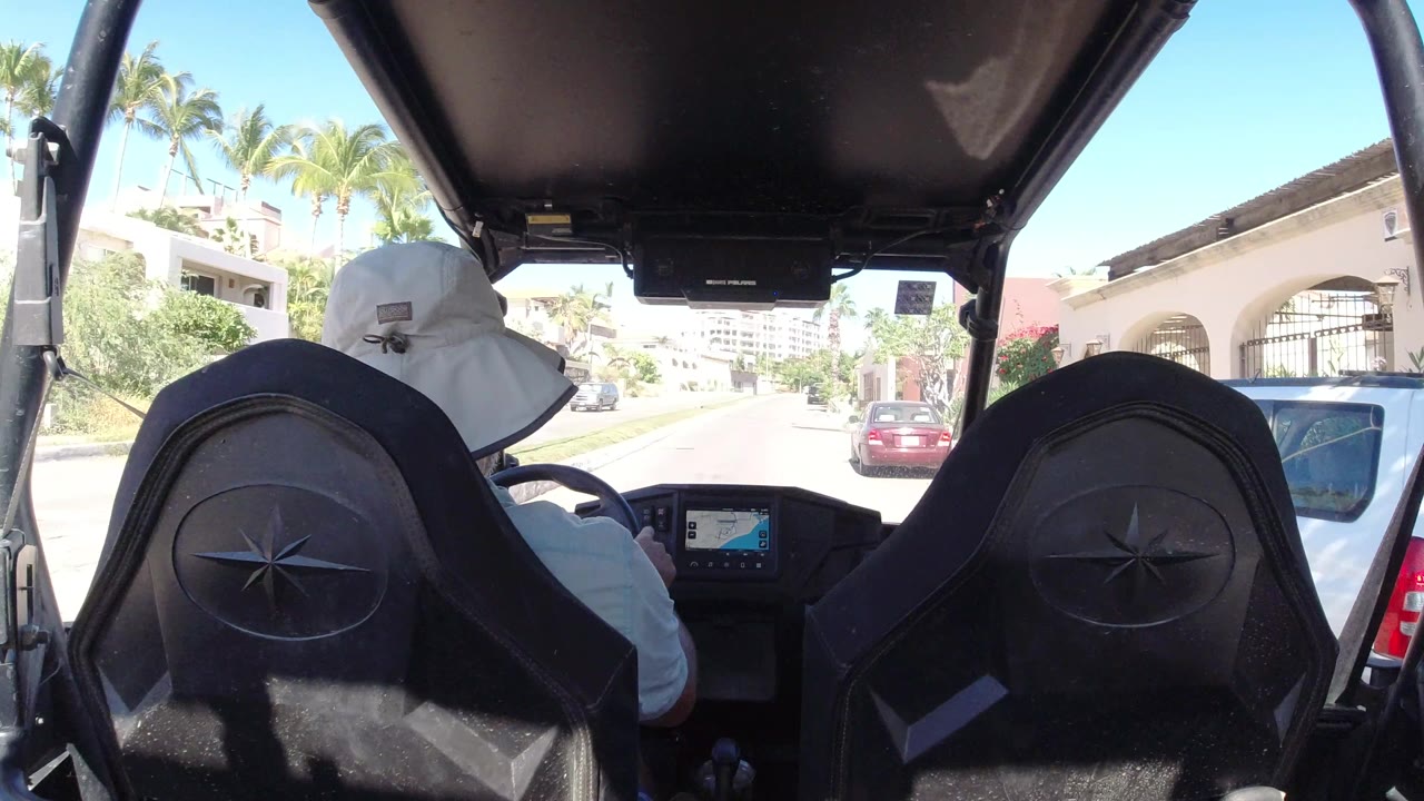 Rear passenger angle from the go-kart in Los Cabos, Mexico, with the driver centered, a parked vehicle close on the right, palms, walls, and bright street glare.