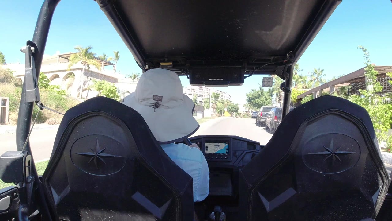 Rear passenger view in Los Cabos, Mexico, with black vehicle seats and roof frame in front of a sunlit residential street and palms.