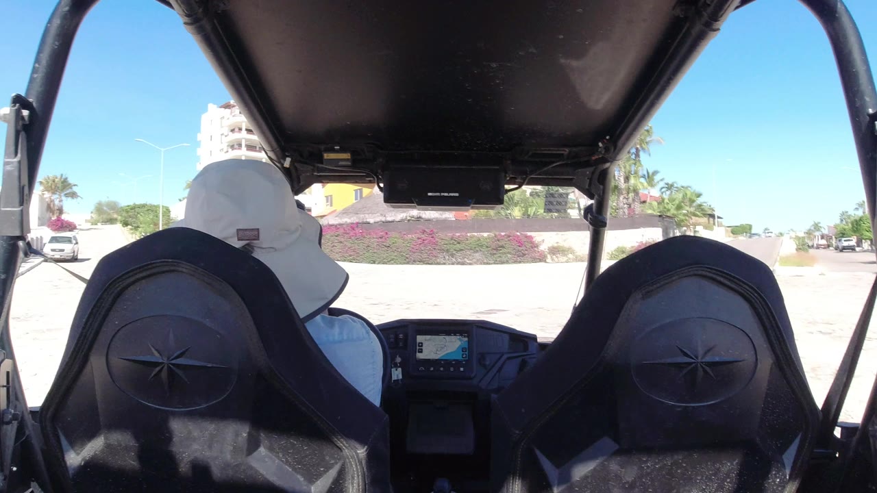 Open vehicle interior in Los Cabos, Mexico, looking past two black seats toward the driver, a dashboard screen, pale pavement, flowering shrubs, parked cars, and low buildings under strong sun.
