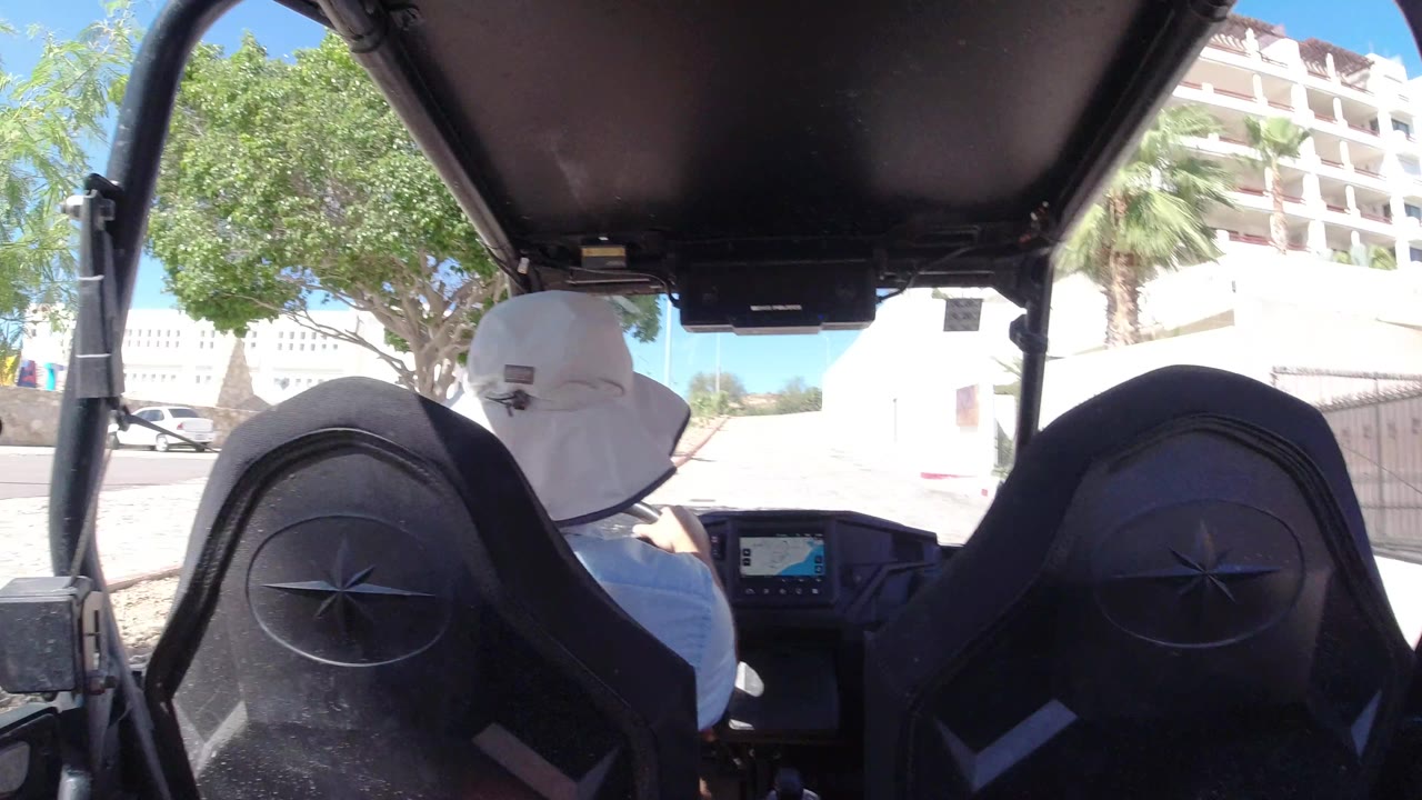 The camera remains behind the driver in the open vehicle as white buildings, a leafy tree, palm fronds, and bright paved ground fill the forward view in Los Cabos, Mexico.